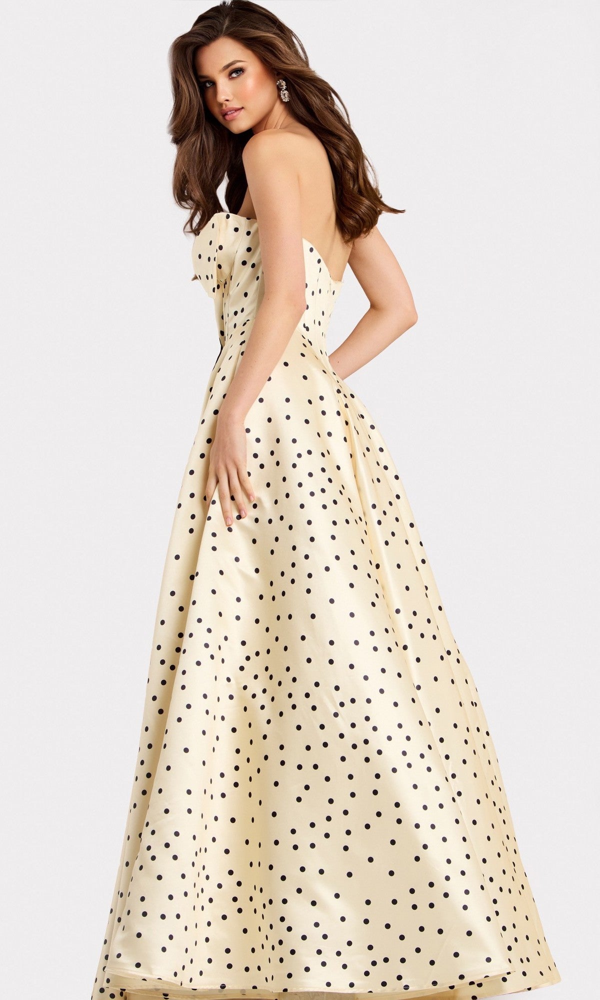 A woman wearing the Jovani 49775, a cream floor-length strapless satin prom dress with black polka dots, stands looking over her shoulder against a plain background.